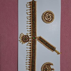 May include: A gold necklace with green and red stones, and two matching gold earrings with green and red stones. The necklace has a long, dangling chain with pearls.