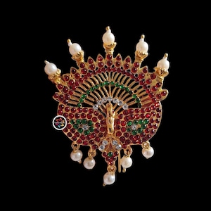 May include: A gold-toned peacock-shaped brooch featuring a fan-like crest adorned with numerous small red and green circular stones. The design includes clear white crystals and several dangling white faux pearls along the top and bottom edges.