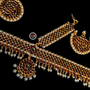 May include: Gold-toned jewelry set featuring intricate designs with red, green, and white accents. The set includes a necklace, earrings, a ring, and a bracelet, all with a delicate, ornate pattern. The jewelry is set against a black background.