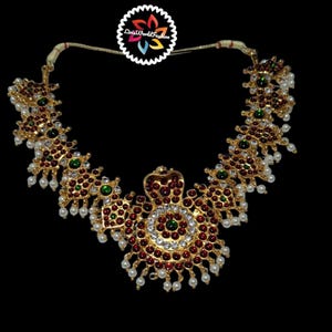 May include: An ornate gold necklace with red and green gemstones, white pearls, and clear crystals. The necklace has a central pendant and a floral design. The background is black.