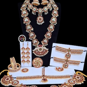 May include: An elaborate Indian jewelry set featuring necklaces, earrings, bracelets, and headpieces. The pieces are adorned with red, green, and white gemstones, set in gold-toned metal. The set includes a variety of designs, including floral and peacock motifs.