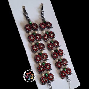 May include: Pair of decorative earrings featuring a cherry design. Each cherry is adorned with red rhinestones and a central clear rhinestone. Green and white accents are also present. The earrings hang from a black chain.