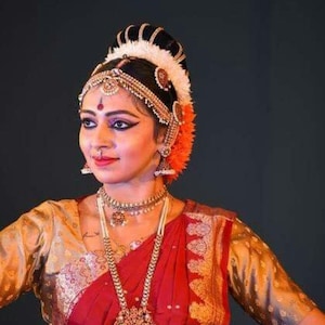 Op de afbeelding: Een vrouw draagt een rode en gouden sari met ingewikkelde borduursels. Ze is versierd met traditionele Indiase sieraden, waaronder een groot hoofddeksel, kettingen en oorbellen. Haar make-up is uitgebreid, met een donkere eyeliner en een rode bindi op haar voorhoofd.