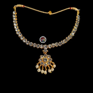 Puede incluir: Un collar de oro con un colgante de piedra blanca y una gota de perla blanca. El collar está hecho de pequeñas piedras blancas y tiene una cadena de oro.