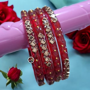 May include: Four red bangles with gold and square crystal accents. The bangles are displayed on a pink surface, with red roses in the background. The jewellery has a textured appearance and is embellished with small gold studs.