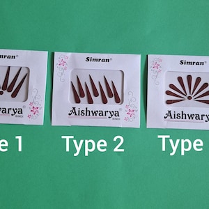 Puede incluir: Tres paquetes de Aishwarya Bindi, cada uno con un diseño diferente. El Tipo 1 presenta un patrón en forma de abanico, el Tipo 2 tiene una serie de puntas y el Tipo 3 muestra un diseño de explosión solar. Los paquetes están sobre un fondo verde.