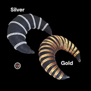 May include: Two crescent-shaped ornaments with black fabric bases. One features silver-toned beaded stripes labeled Silver, and the other features gold-toned sequin stripes labeled Gold. A small circular logo with a colorful flower icon is visible.