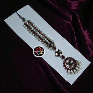 May include: An ornate silver-toned headpiece with red and white accents. The design features a series of connected strands adorned with small white pearls and red stones. A circular pendant with red stones and white pearls hangs at the bottom.