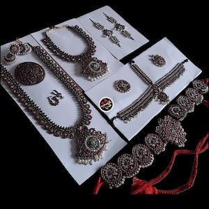 Op de afbeelding: Een assortiment sierlijke sieraden, waaronder kettingen, oorbellen en een ring, gepresenteerd op witte kaarten. De sieraden zijn voorzien van rode en groene edelstenen, met enkele stukken geaccentueerd door kleine witte parels. De achtergrond is zwart.