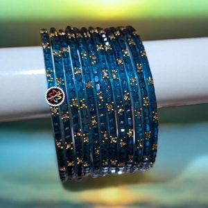May include: A collection of teal blue bangles with gold detailing. The bangles are stacked, revealing a detailed pattern. The jewellery is presented on a white surface with a blurred backdrop.