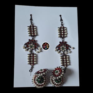 May include: A pair of ornate, dangling earrings and a matching pendant. The earrings and pendant feature a dark metal setting, adorned with red and green gemstones, white pearls, and intricate designs. The jewelry is displayed on a white background.