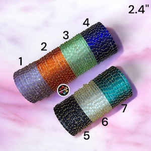 Puede incluir: Se muestran dos pulseras de cuentas, cada una de 6,1 cm de largo. Una pulsera presenta secciones de cuentas lavanda, naranja, verdes y azules. La otra pulsera tiene secciones de cuentas negras, beige y turquesas. Ambas están adornadas con cuentas doradas.