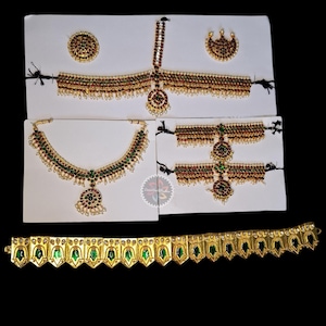 Op de afbeelding: Een assortiment gouden Indiase sieraden, waaronder kettingen, hoofddeksels en oorbellen, versierd met rode, groene en witte edelstenen. De sieraden worden gepresenteerd op witte kaarten tegen een zwarte achtergrond.