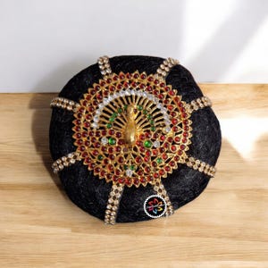 Puede incluir: Un moño negro adornado con una pieza central dorada en forma de pavo real. La pieza central está adornada con piedras preciosas rojas, verdes y transparentes. Acentos dorados con piedras preciosas transparentes rodean el moño.