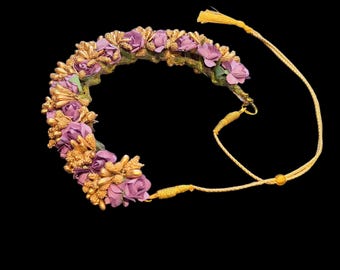 May include: A floral crown featuring purple and gold-colored artificial flowers. The crown is adorned with small, delicate blossoms and gold accents. A gold-colored cord with a tassel is attached to the crown.
