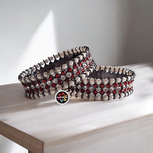 Op de afbeelding: Twee sierlijke, donkergrijze armbanden met ingewikkelde details. Elke armband is versierd met afwisselende rijen witte parels, rode edelstenen en heldere kristallen. De sieraden liggen op een lichtgekleurd houten oppervlak.
