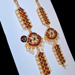 May include: A pair of gold-toned earrings with red and white accents. The earrings feature a combination of red gemstones and white pearls, arranged in a decorative pattern. The design includes a central circular element with a green gemstone.