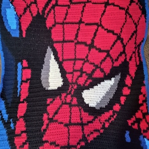 Puede incluir: Una manta de ganchillo con un primer plano de la cara de Spider-Man. La manta es roja, negra y blanca con un borde azul.