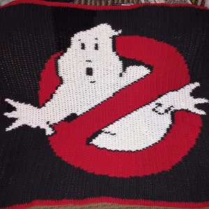 May include: Crocheted blanket featuring the Ghostbusters logo. The logo is red and white with a black background. The logo is a stylised 'no' symbol with a ghost inside.
