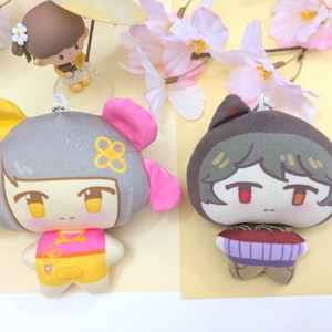 May include: Two plush character keychains. The left plush has grey hair, a yellow and pink bow, and a pink and yellow outfit. The right plush has dark brown hair, cat ears, and a purple and brown striped shirt. Both have small metal ball chains.