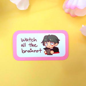 May include: A pink sticker with rounded corners featuring the text "Watch all the brainrot" and an anime-style illustration of a person holding a tablet. The character has dark green hair and is wearing a black jacket.