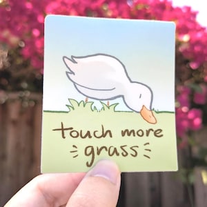 May include: A square sticker featuring a cartoon duck with its beak touching green grass. The sticker has the text "touch more grass" in a handwritten style. The background is a gradient of blue and green.