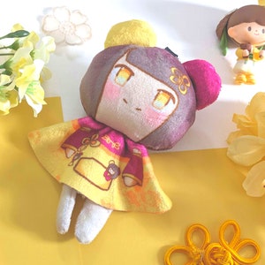 May include: A plush toy doll with brown hair, yellow and pink clothing, and a yellow flower on its head. The doll is sitting on a yellow surface with yellow flowers in the background.