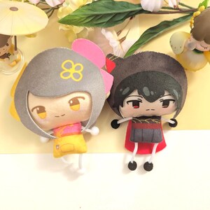 May include: Two plush character dolls with stylized features. The left doll has grey hair with a yellow flower icon, a pink bow, and a yellow and pink outfit. The right doll has black hair, a dark grey hood, and a red cape. Both have thin white limbs.