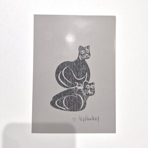 Puede incluir: Una postal gris con un grabado en linóleo en blanco y negro de dos gatos estilizados. Los gatos están apilados, uno encima del otro. La firma del artista está en la parte inferior.