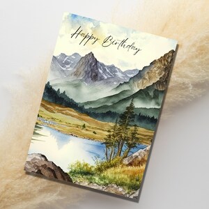 Puede incluir: Una pintura de acuarela de una cordillera con un lago en primer plano. Las monta&ntilde;as est&aacute;n pintadas en tonos de azul, verde y marr&oacute;n. El lago es de color azul claro. El texto "Happy Birthday" est&aacute; escrito en cursiva negra en la parte superior de la tarjeta.