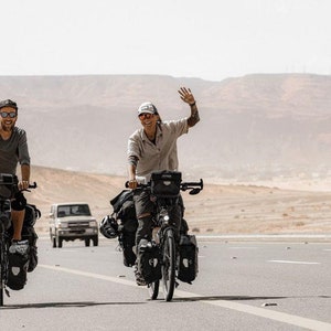 Pode incluir: Duas pessoas andando de bicicleta em uma estrada pavimentada em uma paisagem des&eacute;rtica. Ambos est&atilde;o usando &oacute;culos de sol e sorrindo. A pessoa &agrave; esquerda est&aacute; vestindo uma camiseta cinza e a pessoa &agrave; direita est&aacute; vestindo uma camisa bege. Ambas as bicicletas est&atilde;o carregadas com alforjes.