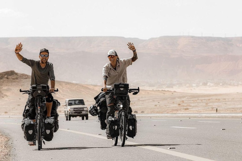 Pode incluir: Duas pessoas andando de bicicleta em uma estrada pavimentada em uma paisagem des&eacute;rtica. Ambos est&atilde;o usando &oacute;culos de sol e sorrindo. A pessoa &agrave; esquerda est&aacute; vestindo uma camiseta cinza e a pessoa &agrave; direita est&aacute; vestindo uma camisa bege. Ambas as bicicletas est&atilde;o carregadas com alforjes.