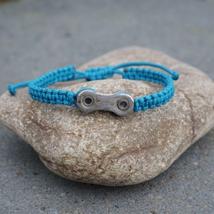 Pode incluir: Uma pulseira tran&ccedil;ada azul turquesa com um fecho de elo de corrente de bicicleta prateado. O fecho tem a palavra "SRAM" gravada.