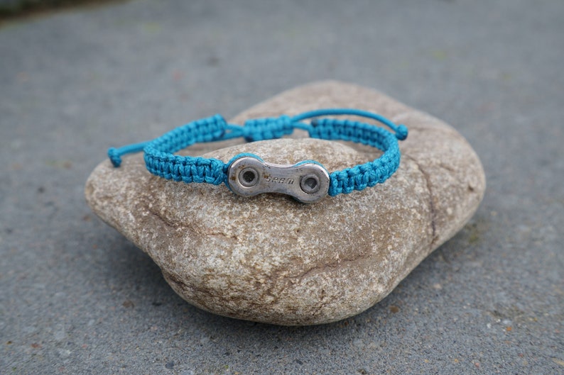 Pode incluir: Uma pulseira tran&ccedil;ada azul turquesa com um fecho de elo de corrente de bicicleta prateado. O fecho tem a palavra "SRAM" gravada.