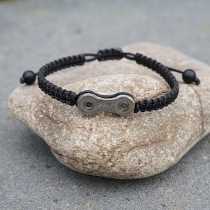 Pode incluir: Um bracelete tran&ccedil;ado preto com um fecho de corrente de bicicleta prateado. O bracelete est&aacute; sobre uma pedra cinzenta.