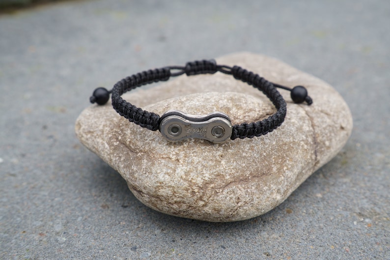 Pode incluir: Um bracelete tran&ccedil;ado preto com um fecho de corrente de bicicleta prateado. O bracelete est&aacute; sobre uma pedra cinzenta.