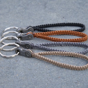 Puede incluir: Cuatro llaveros hechos con un eslabón de cadena de bicicleta de metal y un cordón trenzado. Los cordones son negro, marrón, gris y beige.