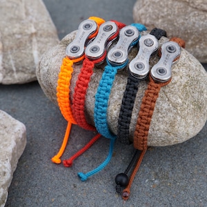 Pode incluir: Cinco pulseiras tran&ccedil;adas coloridas com elos de corrente de bicicleta prateados. As pulseiras est&atilde;o dispostas sobre uma rocha cinza.