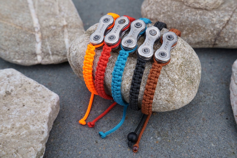 Pode incluir: Cinco pulseiras tran&ccedil;adas coloridas com elos de corrente de bicicleta prateados. As pulseiras est&atilde;o dispostas sobre uma rocha cinza.
