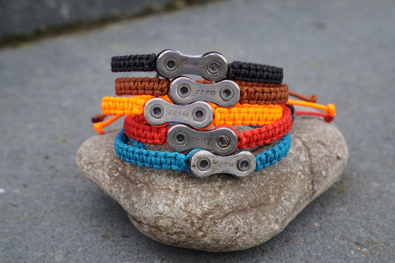 Pode incluir: Cinco pulseiras tran&ccedil;adas coloridas com um fecho de elo de corrente prateado. Cada pulseira tem a palavra "SRAM" gravada no fecho.