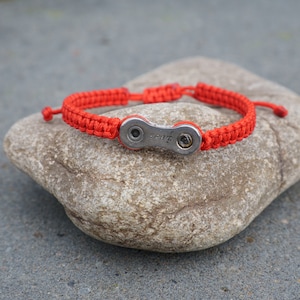 Pode incluir: Um bracelete tran&ccedil;ado vermelho com um fecho de elo de corrente de bicicleta prateado. O bracelete est&aacute; sobre uma pedra cinzenta.
