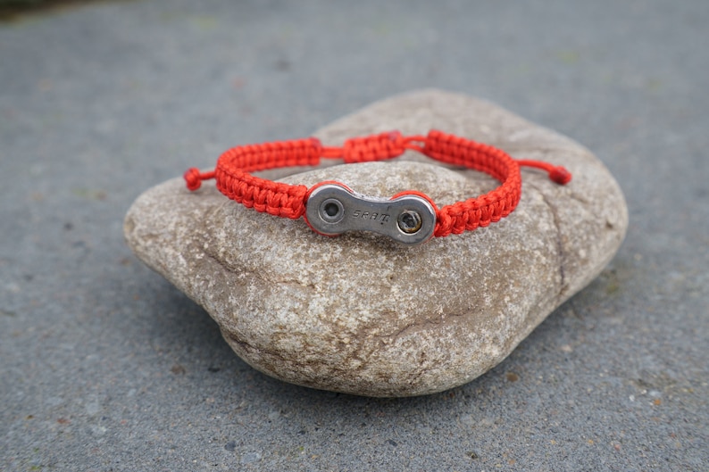 Pode incluir: Um bracelete tran&ccedil;ado vermelho com um fecho de elo de corrente de bicicleta prateado. O bracelete est&aacute; sobre uma pedra cinzenta.