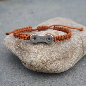 Pode incluir: Uma pulseira tran&ccedil;ada marrom com um charme de elo de corrente de bicicleta prateado. O charme tem a palavra "SRAM" gravada.