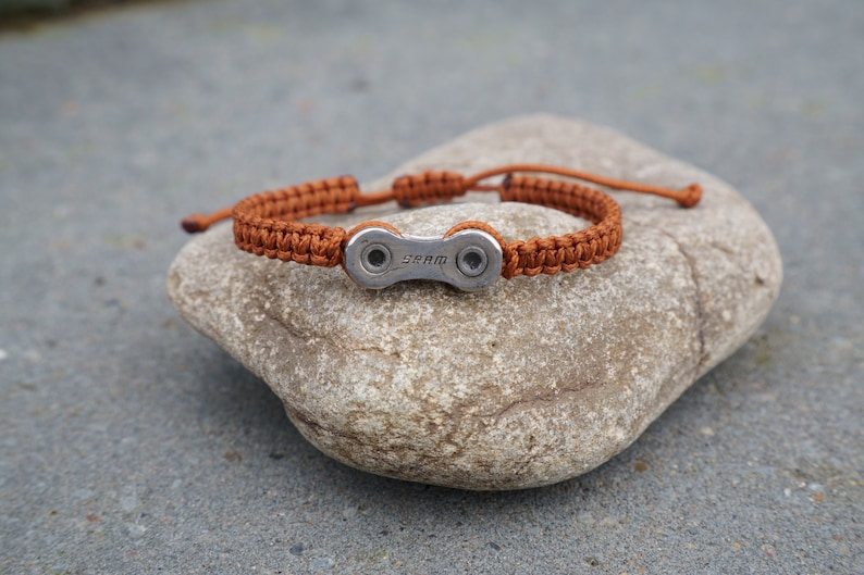 Pode incluir: Uma pulseira tran&ccedil;ada marrom com um charme de elo de corrente de bicicleta prateado. O charme tem a palavra "SRAM" gravada.