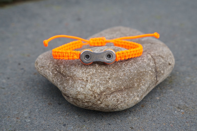 Pode incluir: Pulseira tran&ccedil;ada laranja com um fecho de elo de corrente de bicicleta prateado. O fecho tem a palavra "SRAM" gravada.