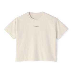 May include: A cream-coloured t-shirt with short sleeves and a crew neck. The text "TINY RIOT" is printed in a small, dark font centered on the chest. The shirt is made of a soft, opaque fabric.