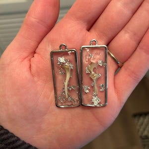 May include: Two rectangular pendants with silver frames, each containing a bone-like structure and silver flakes suspended in clear resin. The pendants are displayed on a person's open palm.