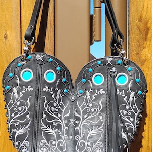 Op de afbeelding: Zwarte leren handtas met witte bloemenborduurwerk, turkooizen accenten en een decoratieve zilveren gesp. Dubbel compartiment ontwerp, zwarte riem en franjes. Een stijlvol accessoire.