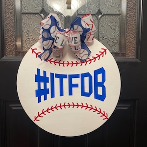 Puede incluir: Colgador de puerta redondo de madera blanco con forma de pelota de béisbol con costuras rojas. Un lazo azul y blanco con temática de béisbol está sujeto a la parte superior. Las palabras "#ITFDB" están escritas en letras azules en la parte delantera del colgador de puerta.