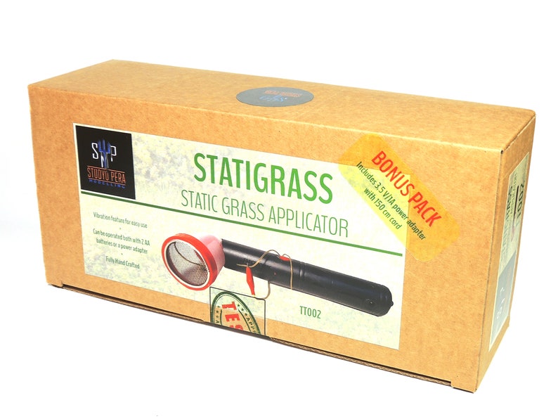 K&ouml;nnte beinhalten: Ein brauner Karton mit einem Etikett, auf dem "Statigrass Static Grass Applicator" steht und ein Bild des Produkts abgebildet ist. Das Produkt ist ein schwarzes Handger&auml;t mit einem roten und silbernen Netz oben. Die Box enth&auml;lt au&szlig;erdem ein Bonus-Paket mit einem 3,5 V/1A Netzteil und einem 150 cm langen Kabel.
